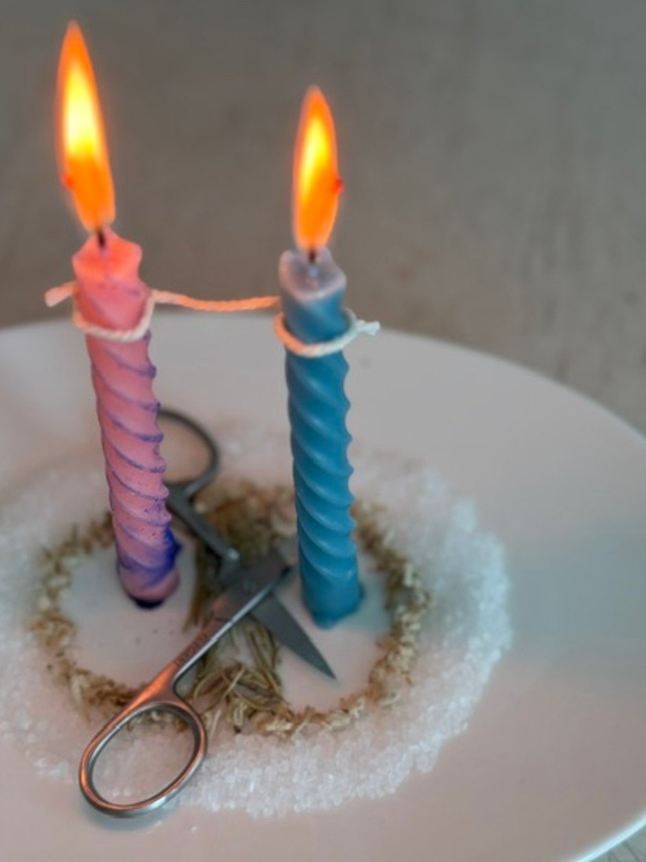 Two twisted candles, one pink and one blue, tied with a cord on a plate of salt and herbs, with scissors between them