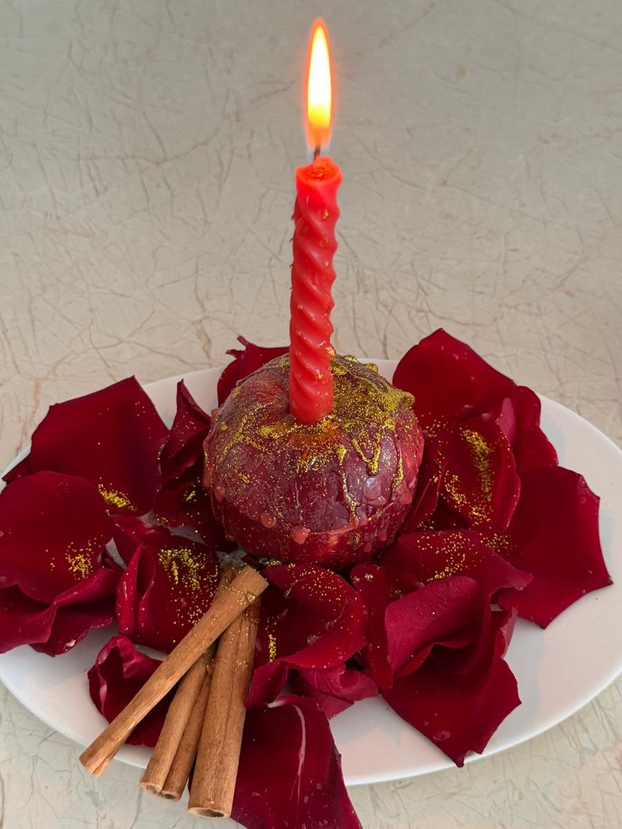 A red candle burning on an apple covered in honey and red rose petals, with cinnamon sticks beside it