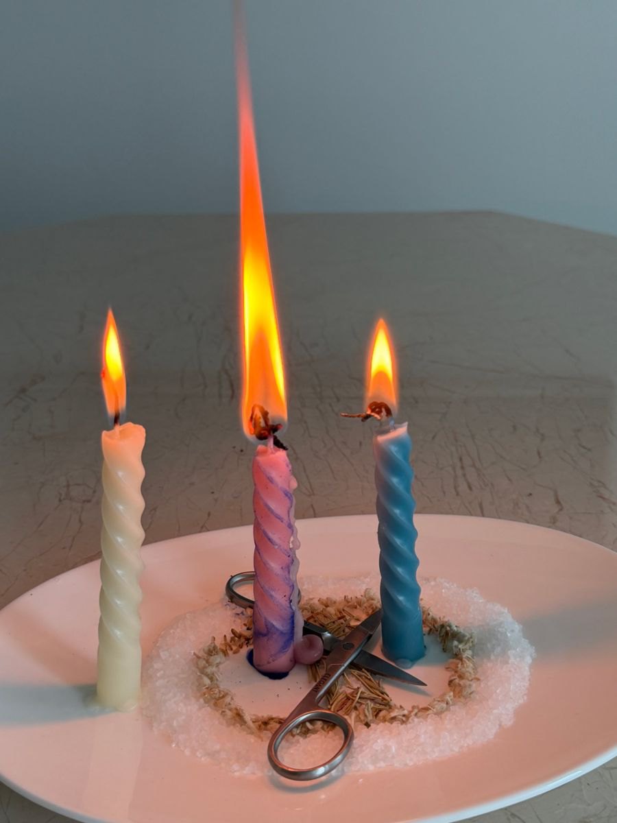 Three twisted candles burning on a plate of salt and herbs, with scissors laid between them